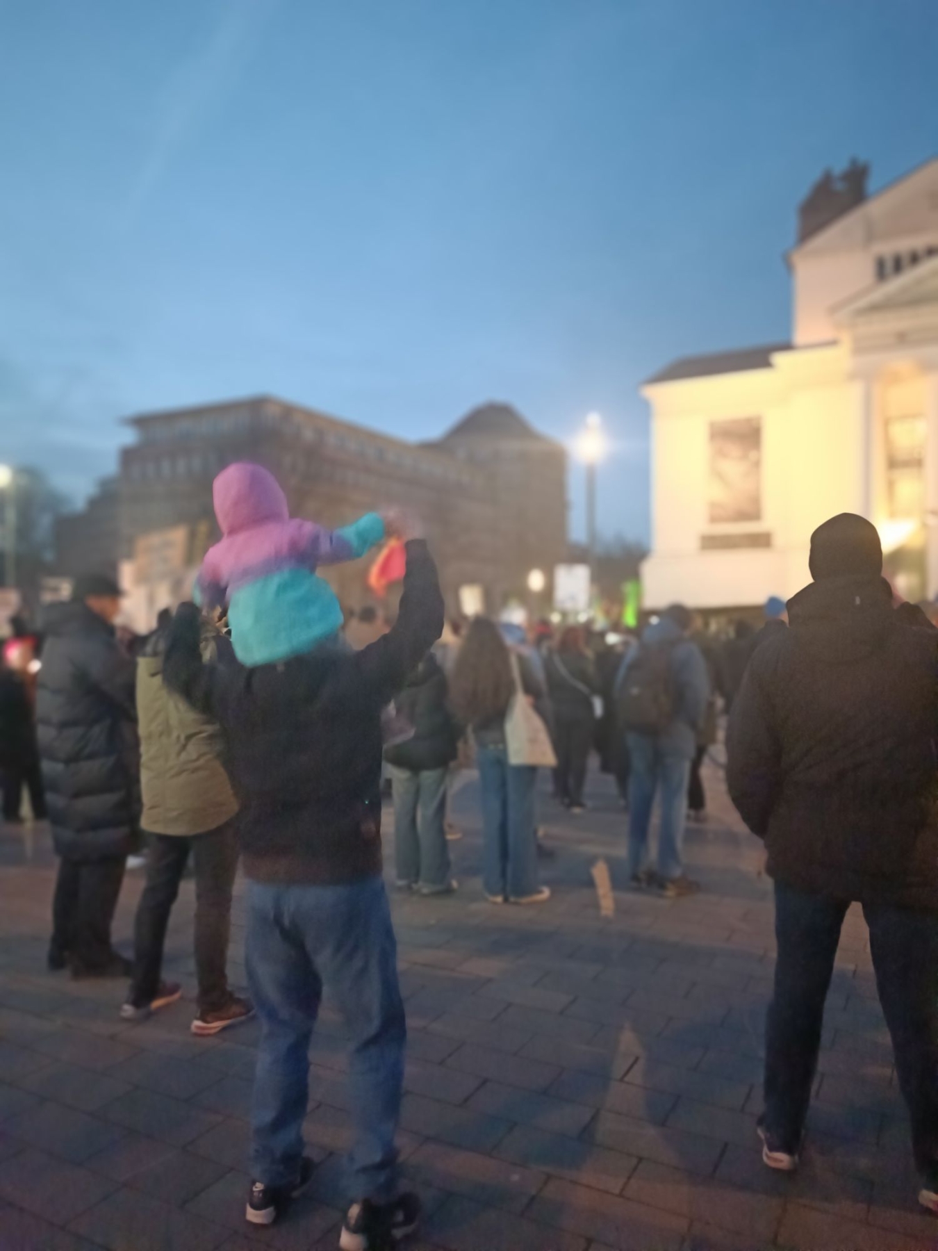 Ein Erwachsener trägt ein Kind auf den Schultern während einer Versammlung bei Dämmerung.
