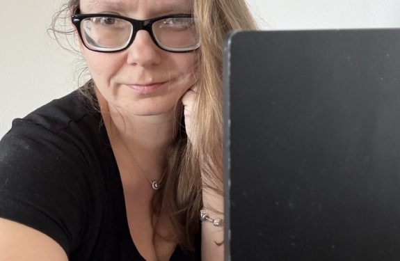 Frau mit langen Haaren und Brille, die an einem Laptop sitzt und in die Kamera schaut.