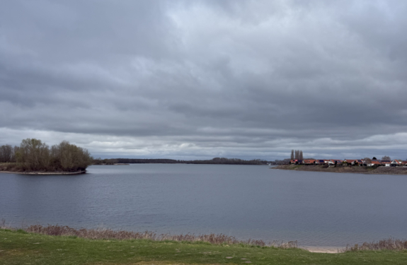 Ein breiter See liegt unter einem dichten, grauen Wolkenhimmel; am Ufer stehen Bäume und eine Reihe rotdachiger Häuser.