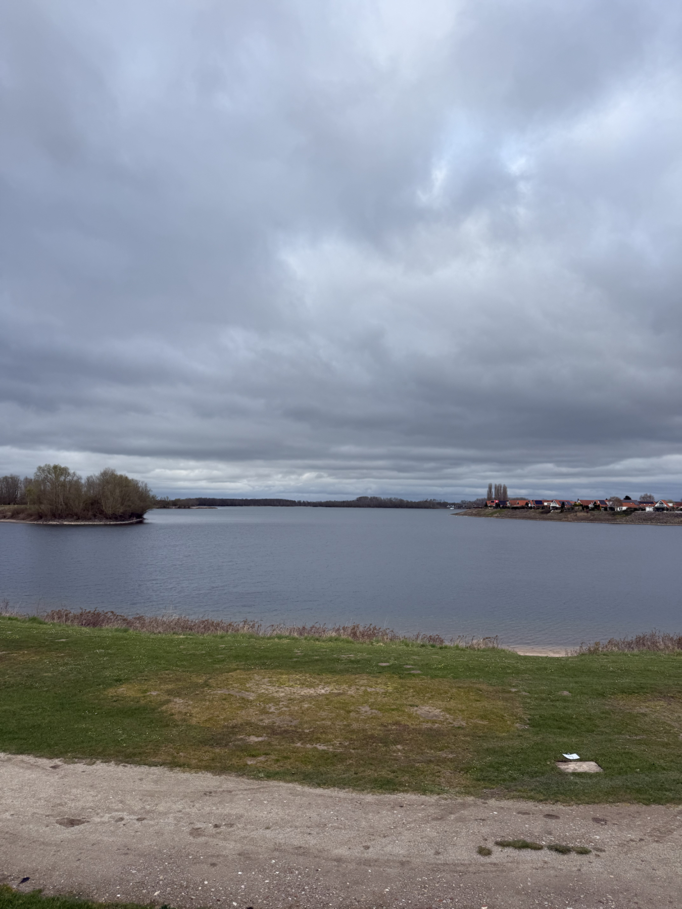Ein breiter See liegt unter einem dichten, grauen Wolkenhimmel; am Ufer stehen Bäume und eine Reihe rotdachiger Häuser.