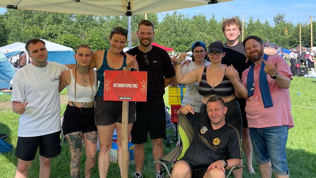 Eine Gruppe von Festivalbesuchern steht und sitzt unter einem weißen Pavillon auf einer grünen Wiese, während ein rotes Schild in der Mitte hängt.