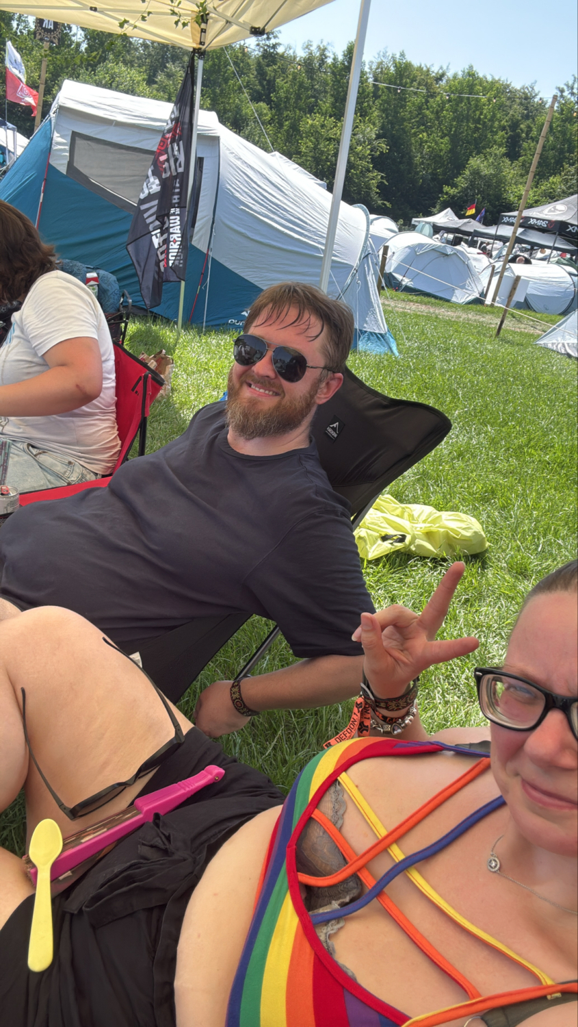 Mehrere Freunde sitzen bei Sonnenschein auf einer Wiese zwischen Festivalzelten, während ein Mann mit Sonnenbrille entspannt im Campingstuhl lehnt.