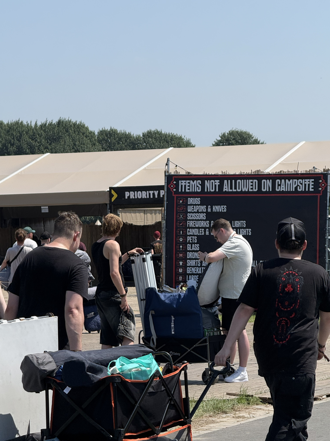 Vor einem Campingbereich stehen Reisende mit Gepäck, während ein großes Schild die auf dem Campsite verbotenen Gegenstände auflistet.