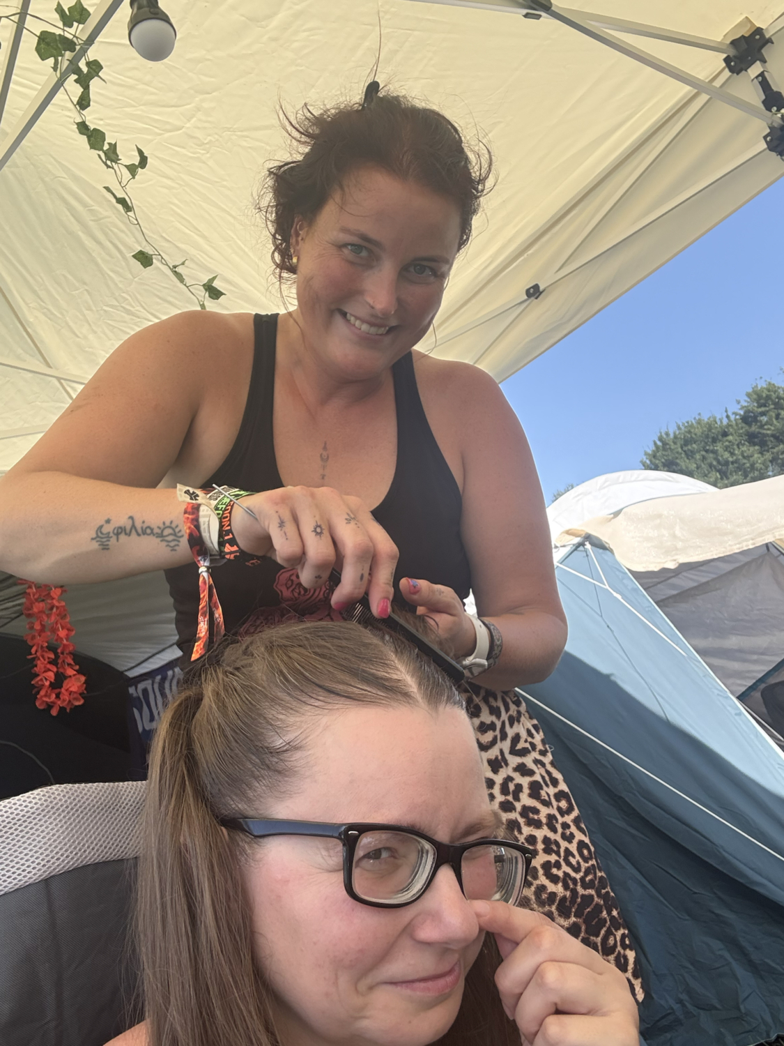 Zwei Frauen sitzen im Freien unter einem hellen Zelt, während die eine der anderen die Haare hochsteckt und beide entspannt lächeln bei Sonnenschein.