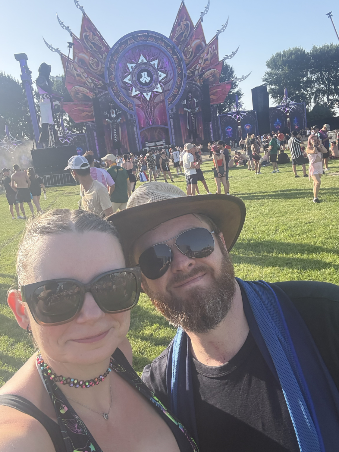 Ein lächelndes Paar macht ein Selfie vor einer großen Bühne auf einem Musikfestival im Freien