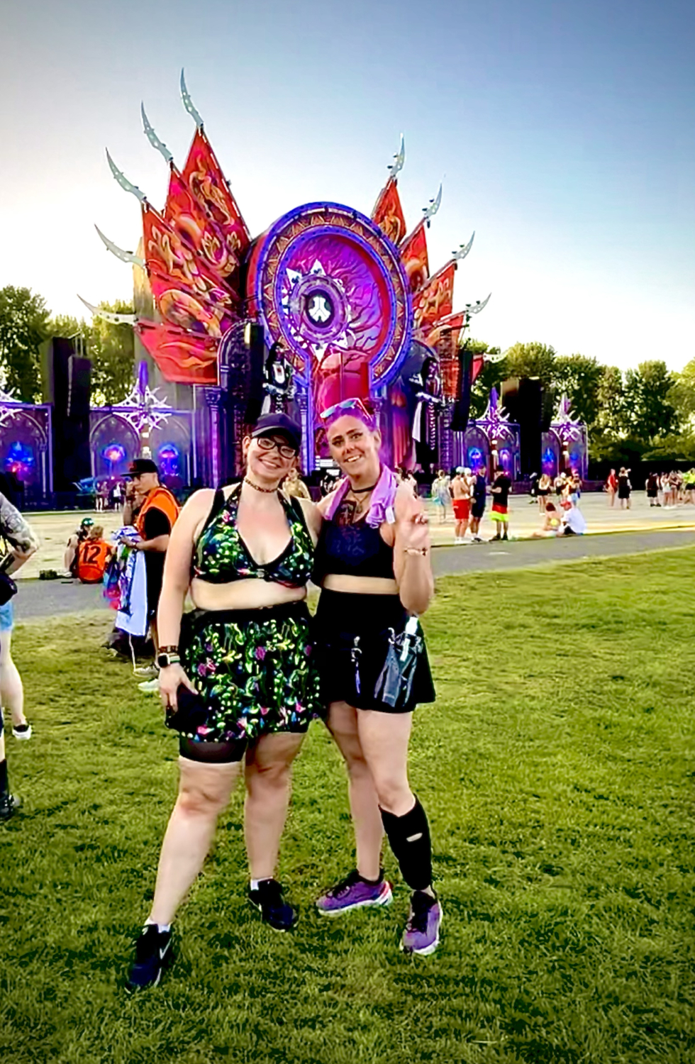 Zwei Frauen lächeln vor einer farbenfrohen Festivalbühne mit lila und roten Ornamenten auf einer grünen Wiese, während Besucher im Hintergrund stehen.