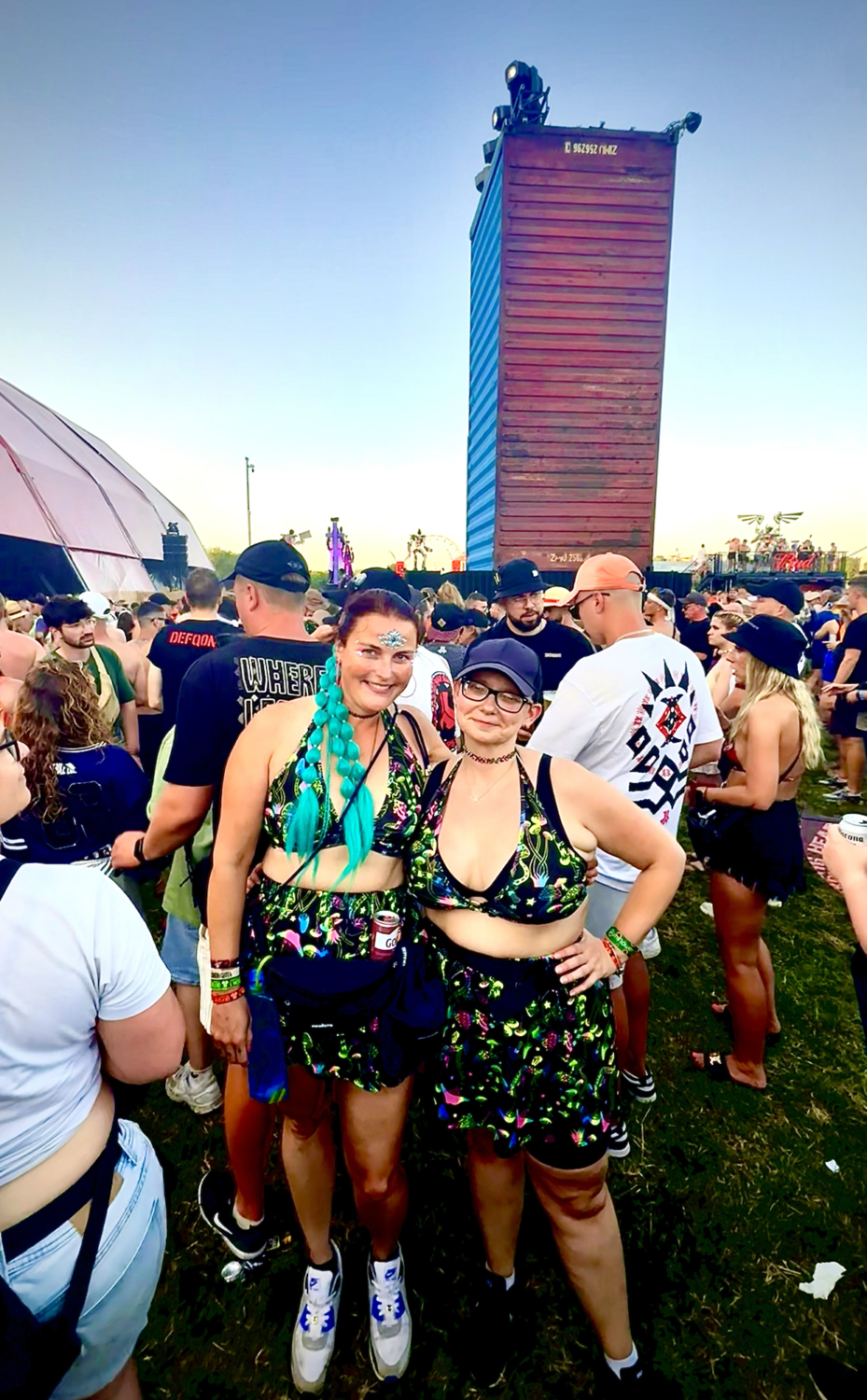 Zwei Frauen posieren auf einer Wiese vor einer bunten Festivalbühne mit leuchtenden Ornamenten und einem klaren Himmel im Hintergrund bei Tageslicht.