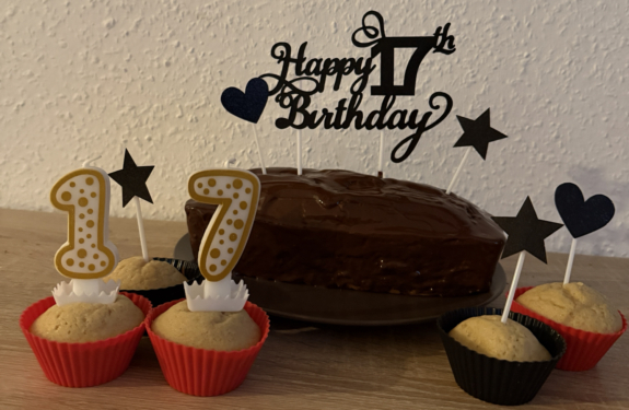 Ein Schokoladenkuchen mit Muffins, Kerzen mit der Zahl 17 sowie schwarzen Sternen und Herzen auf einem Geburtstagstisch.