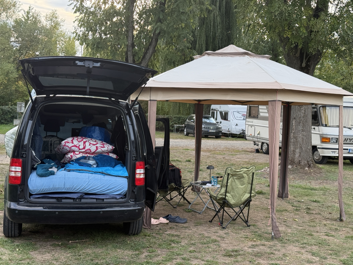 Auf einem Campingplatz steht ein dunkler Van mit ge&ouml;ffneter Heckklappe und Bettzeug neben einem gro&szlig;en Vorzelt mit Campingst&uuml;hlen.