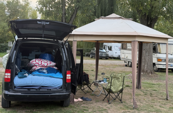 Auf einem Campingplatz steht ein dunkler Van mit geöffneter Heckklappe und Bettzeug neben einem großen Vorzelt mit Campingstühlen.
