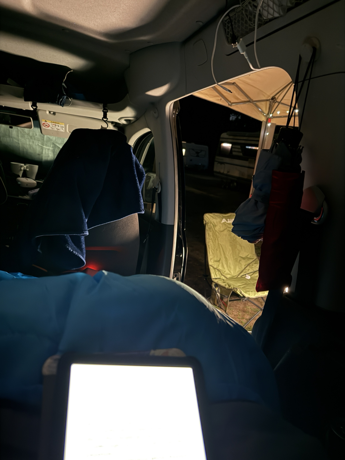 Im ge&ouml;ffneten Campervan liegt ein blaues Bettzeug; drau&szlig;en stehen bei Nacht ein gelber Klappstuhl und ein Sonnendach.