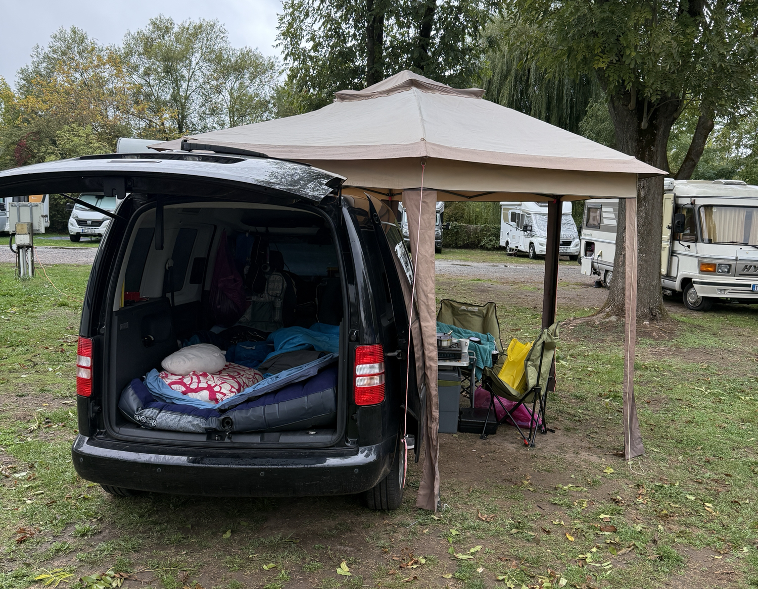 Auf einem Campingplatz steht ein schwarzer Van mit offenem Kofferraum unter einem gro&szlig;en beigen Vorzelt; Matratze und Bettzeug liegen darin.