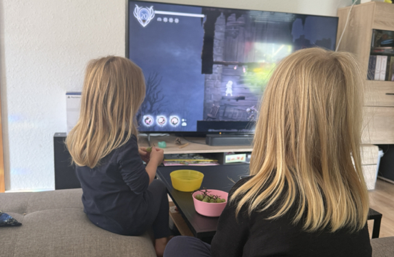 Zwei Kinder sitzen auf dem Sofa und schauen gemeinsam ein Fantasy-Spiel auf dem Fernseher, während vor ihnen Snacks stehen.