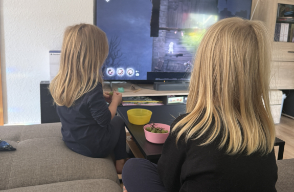 Zwei Kinder sitzen auf dem Sofa und schauen gemeinsam ein Fantasy-Spiel auf dem Fernseher, während vor ihnen Snacks stehen.