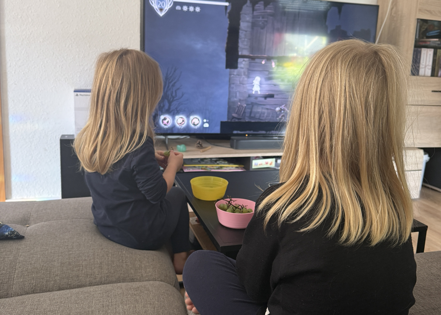 Zwei Kinder sitzen auf dem Sofa und schauen gemeinsam ein Fantasy-Spiel auf dem Fernseher, während vor ihnen Snacks stehen.