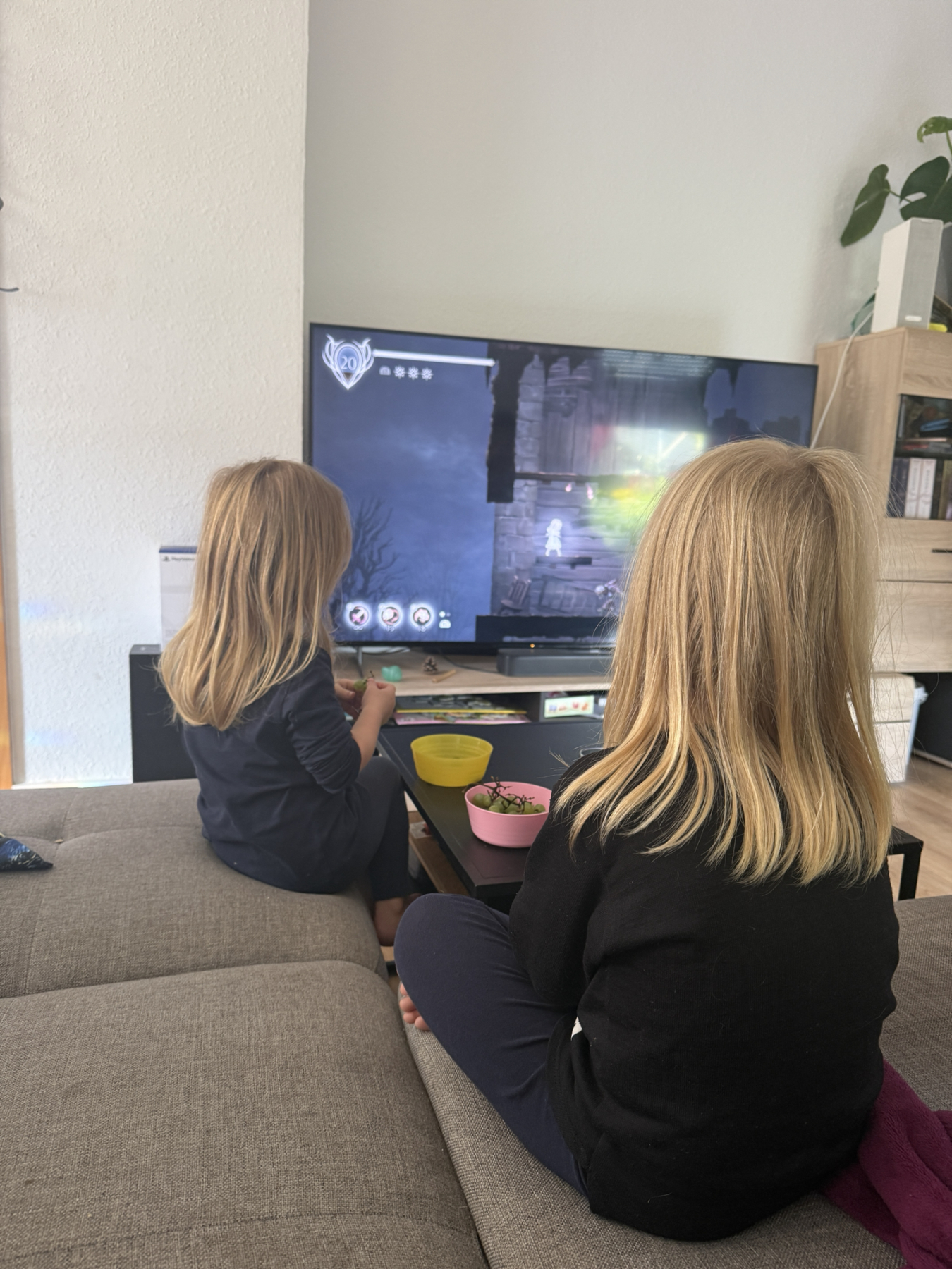 Zwei Kinder sitzen auf dem Sofa und schauen gemeinsam ein Fantasy-Spiel auf dem Fernseher, während vor ihnen Snacks stehen.