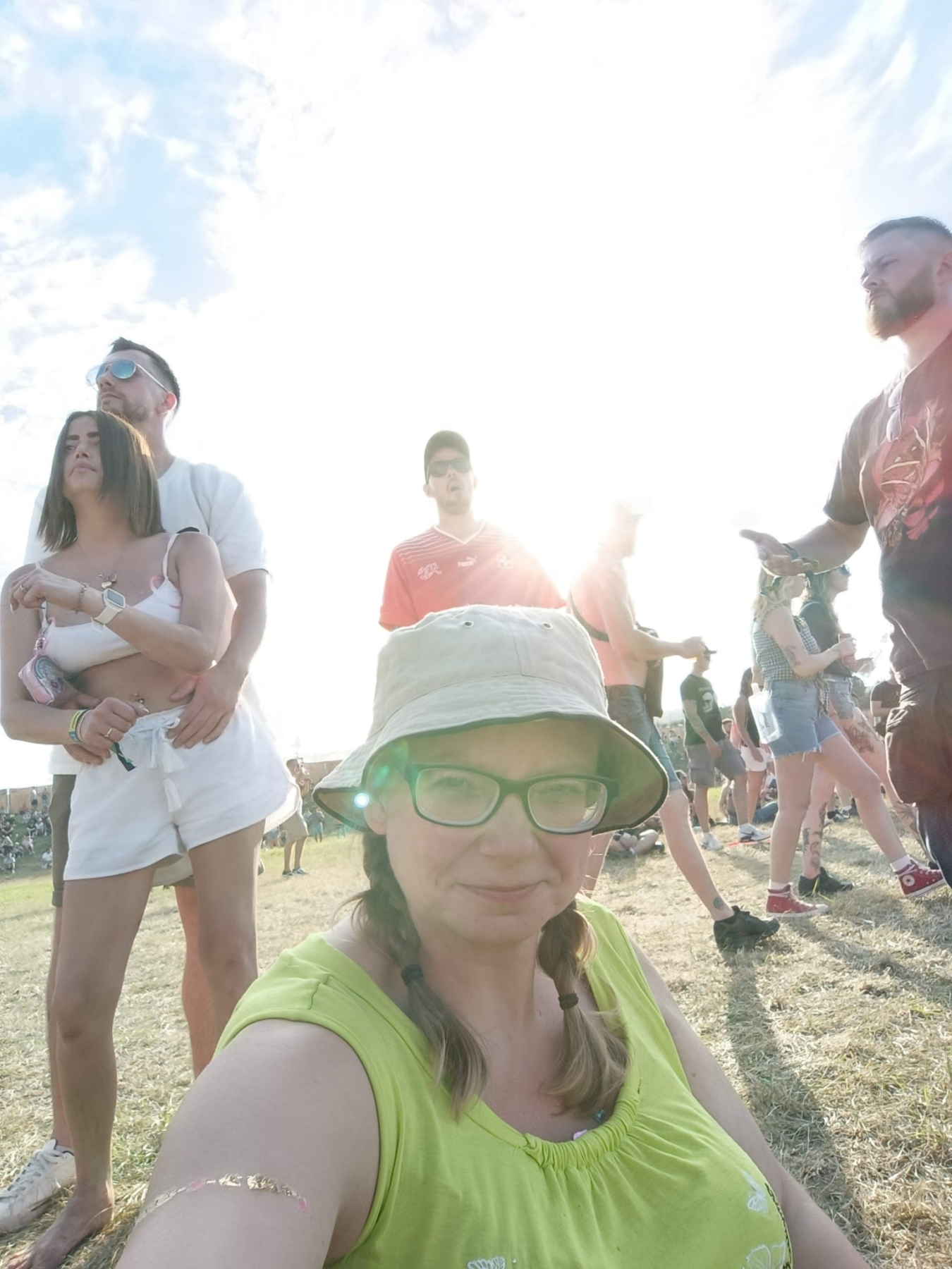 Eine Frau mit Brille und Hut sitzt auf dem Boden während eines Festivals, umgeben von Menschen.