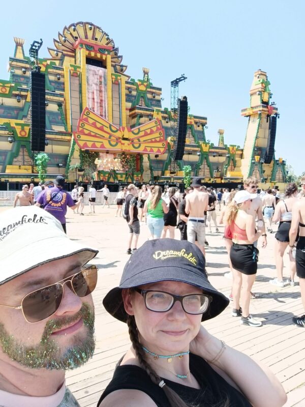Ein Selfie von mir und meinen Mann auf dem Festival Dreamfields. Im Vordergrund stehen mein Mann, er trägt Vollbart, eine Sonnenbrille und einen weißen Schlapphut. Rechts daneben stehe ich, mit schwarzen Schlapphut und trage eine Brille. Hinter uns viele Festivalbesucher vor der Mainstage. Diese ist im Stil der Azteken gestaltet.