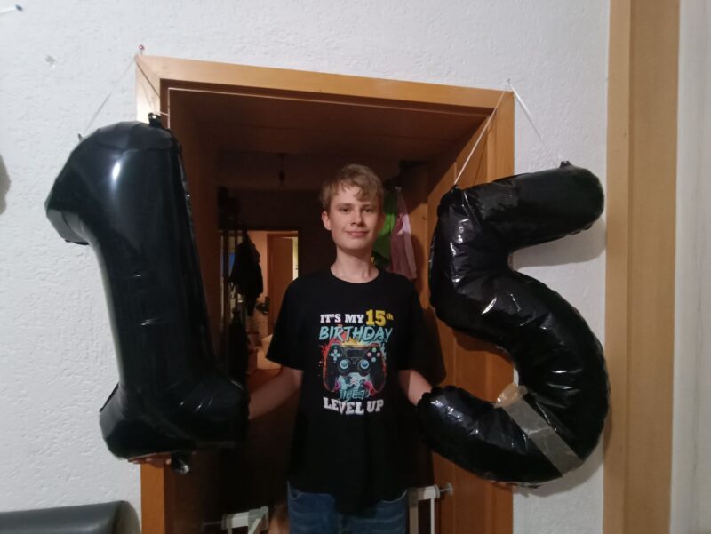 Auf dem Bild steht ein Junge von 15 Jahren, er trägt ein Shirt mit der Aufschrift "It´s my 15. Birthday. Level Up.". Er hält zwei Folienballons in den Händen, eine 1 und eine 5.