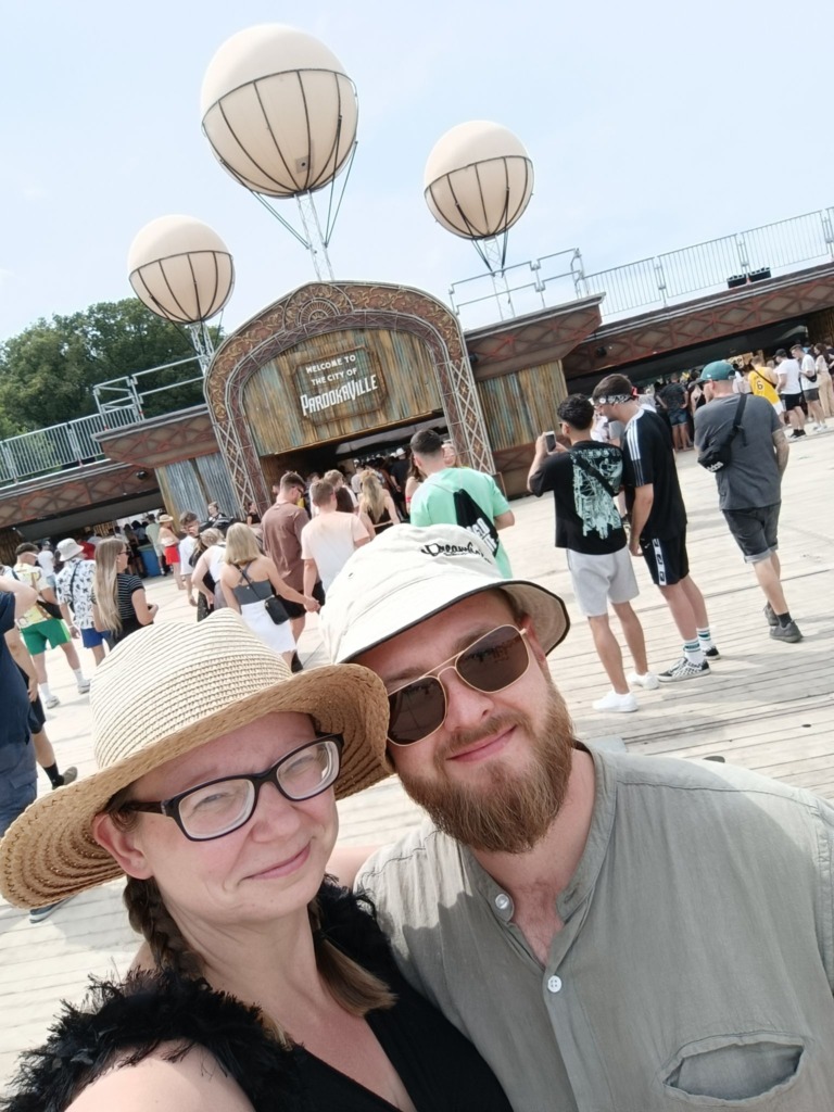Ein Paar mit Sonnenh&uuml;ten posiert vor einem Festivalgel&auml;nde mit Menschenmenge im Hintergrund.
